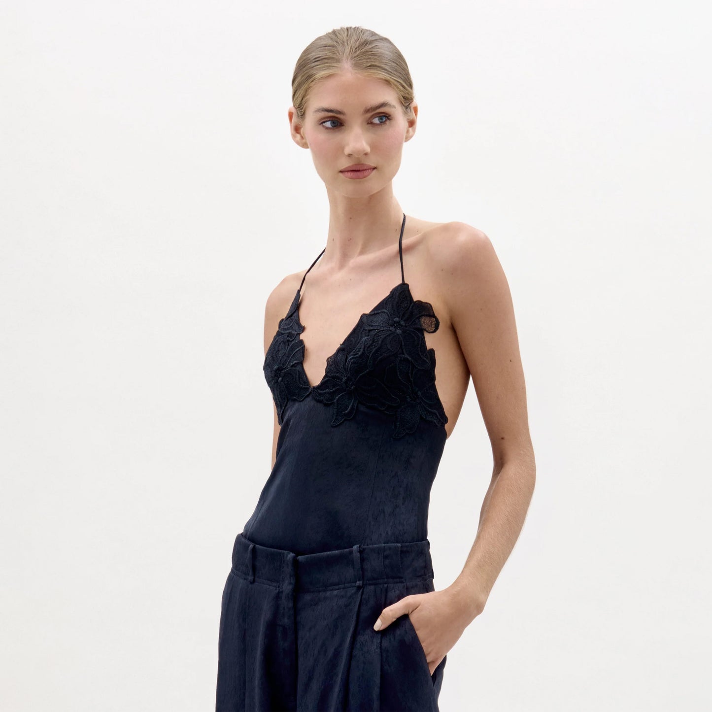A woman with blonde hair tied back wears the Diora Top, a black halter-neck style with floral lace applique, paired with dark high-waisted pants. She poses with one hand in her pocket against a plain white background.