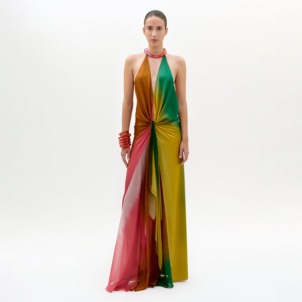 Against a plain white background, a person models the Dixon Dress—a vibrant silk column gown with pink, red, orange, green, and yellow panels gathered at the waist—paired with chunky red bracelets and a necklace.