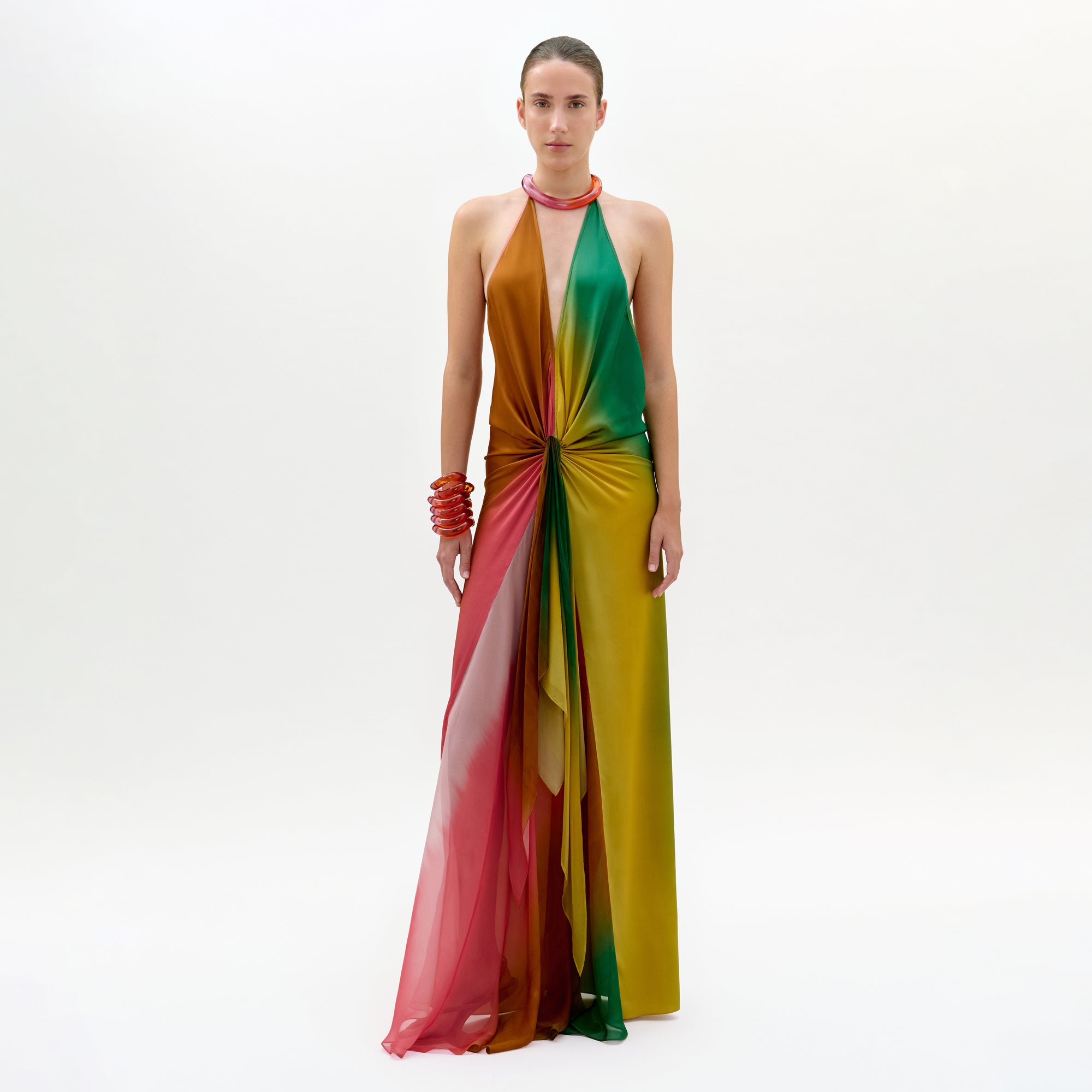 Against a plain white background, a person models the Dixon Dress—a vibrant silk column gown with pink, red, orange, green, and yellow panels gathered at the waist—paired with chunky red bracelets and a necklace.