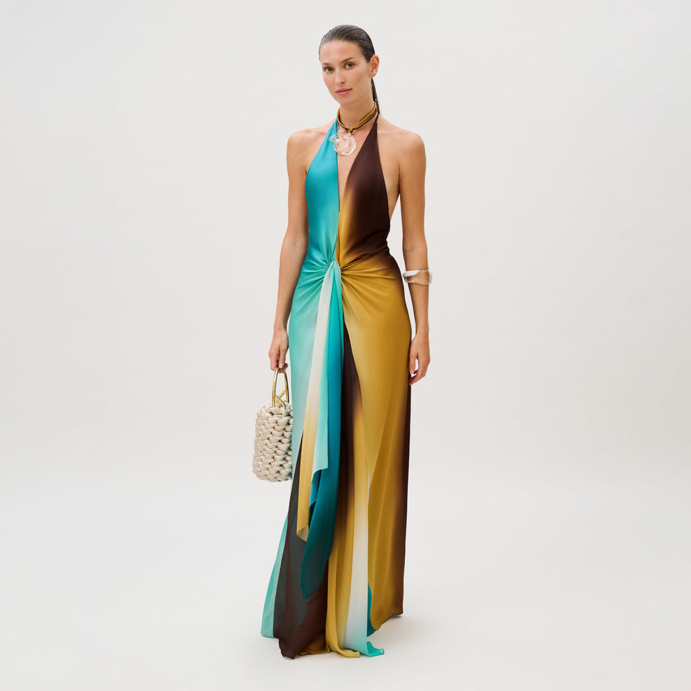 A model wears the Dixon Dress, a floor-length silk chiffon halter-neck gown featuring a color-blocked design of teal, brown, and yellow, with a draped knotted detail at the waist and a woven handbag.