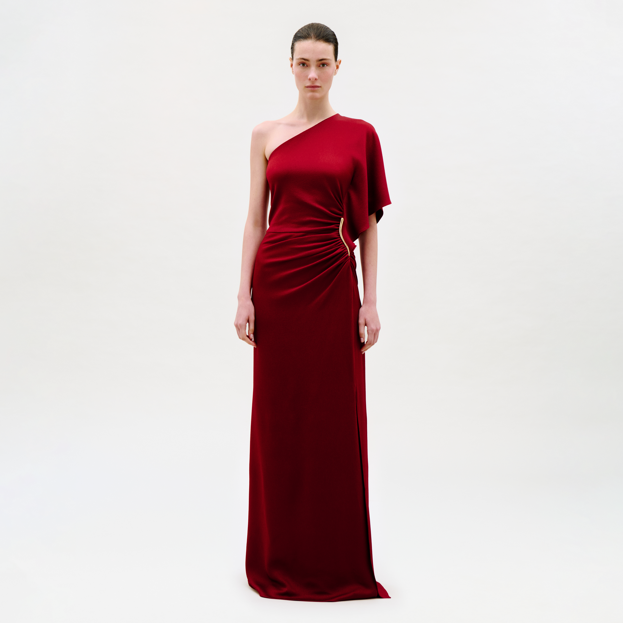 A woman stands against a plain light background, wearing the Doli Dress—a floor-length, one-shoulder dark red gown featuring a gathered waist detail and a single short sleeve.