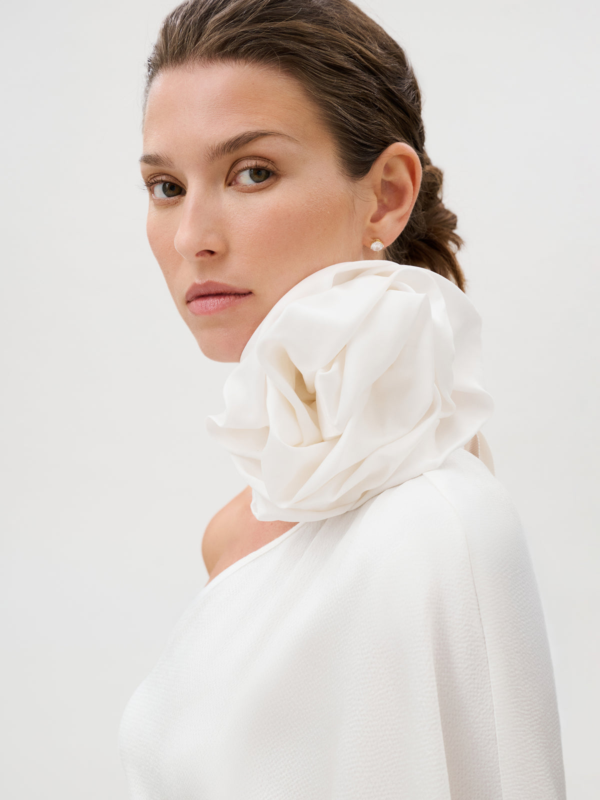 A woman wearing the Doli Dress—a white, off-shoulder piece featuring a large, ruffled collar—gazes confidently at the camera against a plain, light background. Her hair is pulled back to highlight the elegant garment, which is expertly crafted from hammered satin in Italy, adding an extra layer of sophistication to her poised presence.