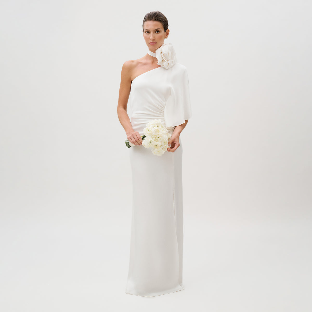 A woman in the Doli Dress—a white, one-shoulder hammered satin gown featuring a large flower detail on the shoulder—holds a small bouquet of white flowers while standing against a plain white background. This elegant dress is made in Italy.