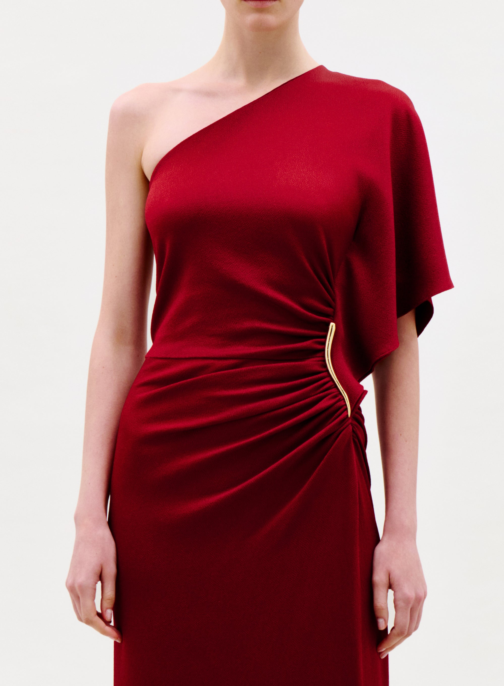 A woman models the Doli Dress, featuring an elegant one-shoulder design in deep red with ruched detailing and a gold waist accent. Her face is not visible, set against a plain white background.
