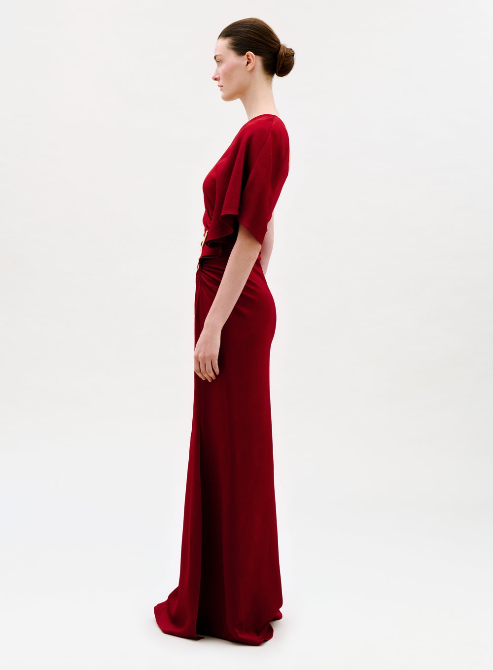 A woman stands in profile against a plain, light background, wearing the floor-length, fitted Doli Dress in red with short sleeves. Her hair is styled in a neat bun and her arms rest naturally by her sides.