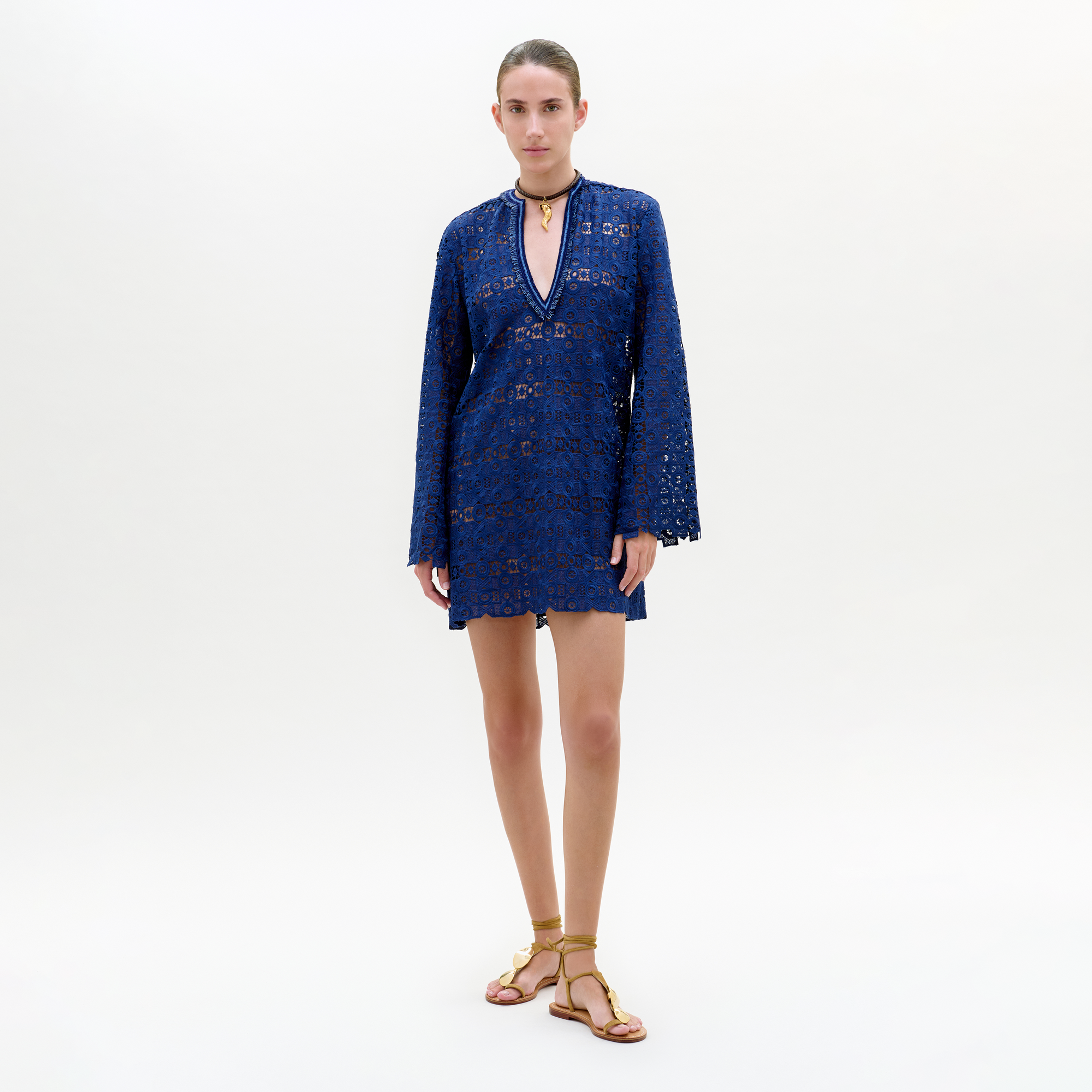 A person stands against a plain white background wearing the Dorenia Dress, a dark blue, long-sleeve mini day dress made from Italian guipure fabric with a deep V-neckline, paired with a gold necklace and flat gold sandals.