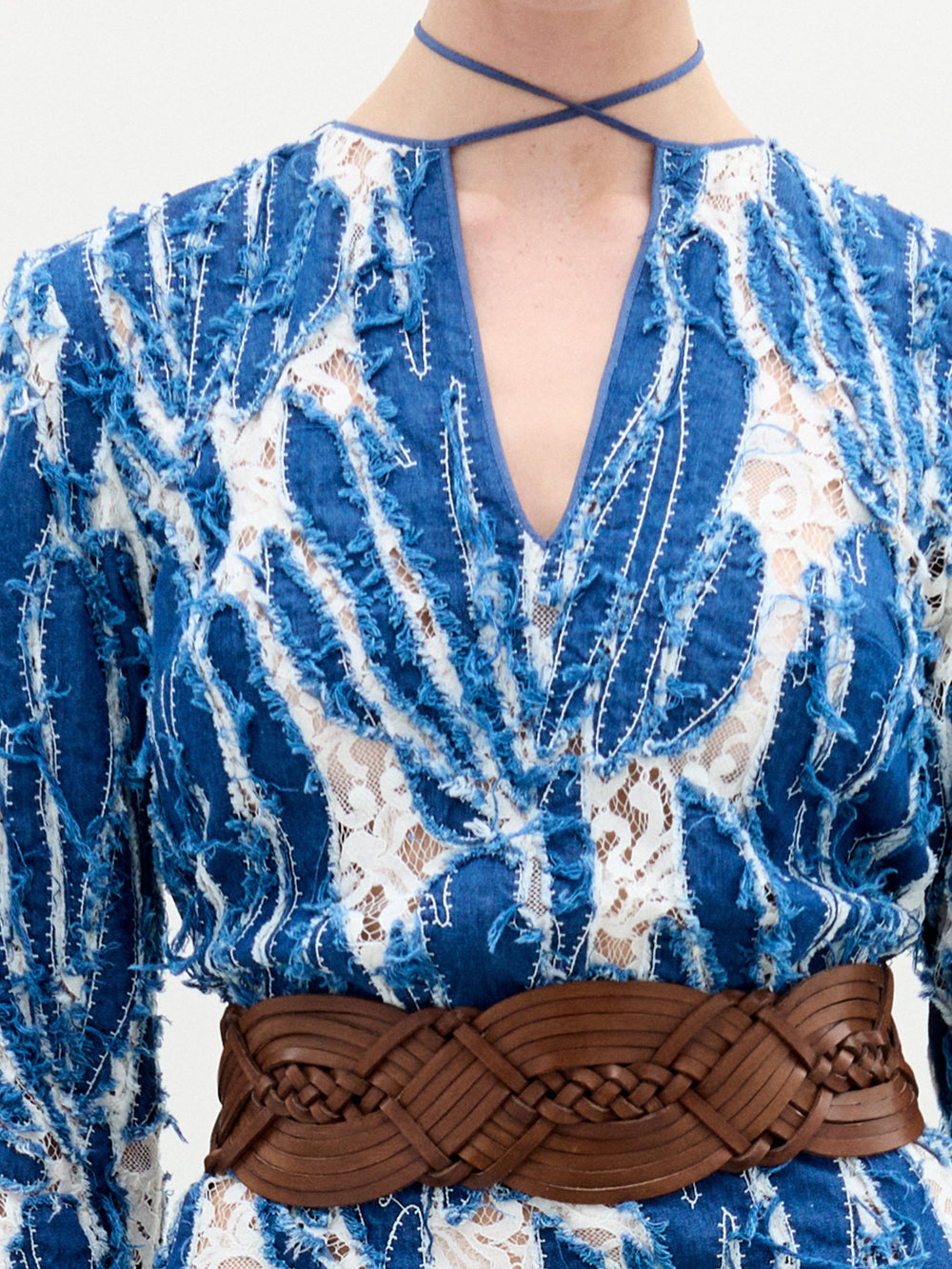 A person stands against a plain background wearing the Ebba Dress, a long blue and white patterned dress with a brown belt, high neckline, and subtle criss-cross detailing that enhances its elegant column silhouette.