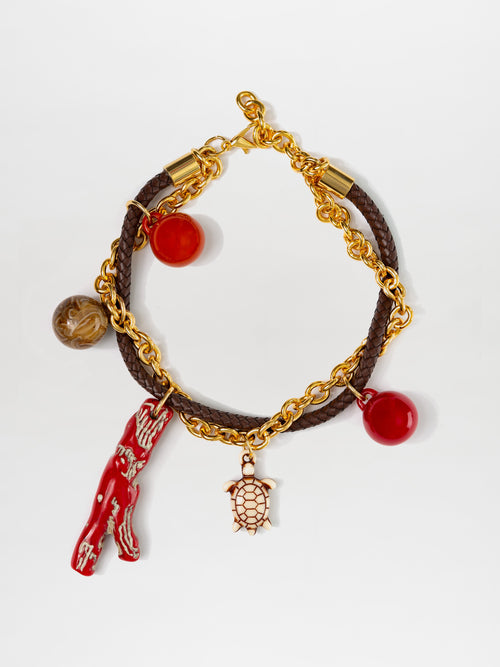 The Eira Necklace features a gold chain and brown braided cord with metallic accents, adorned with red and brown multicolor charms, including a coral-shaped charm and a turtle-shaped charm against a white background.