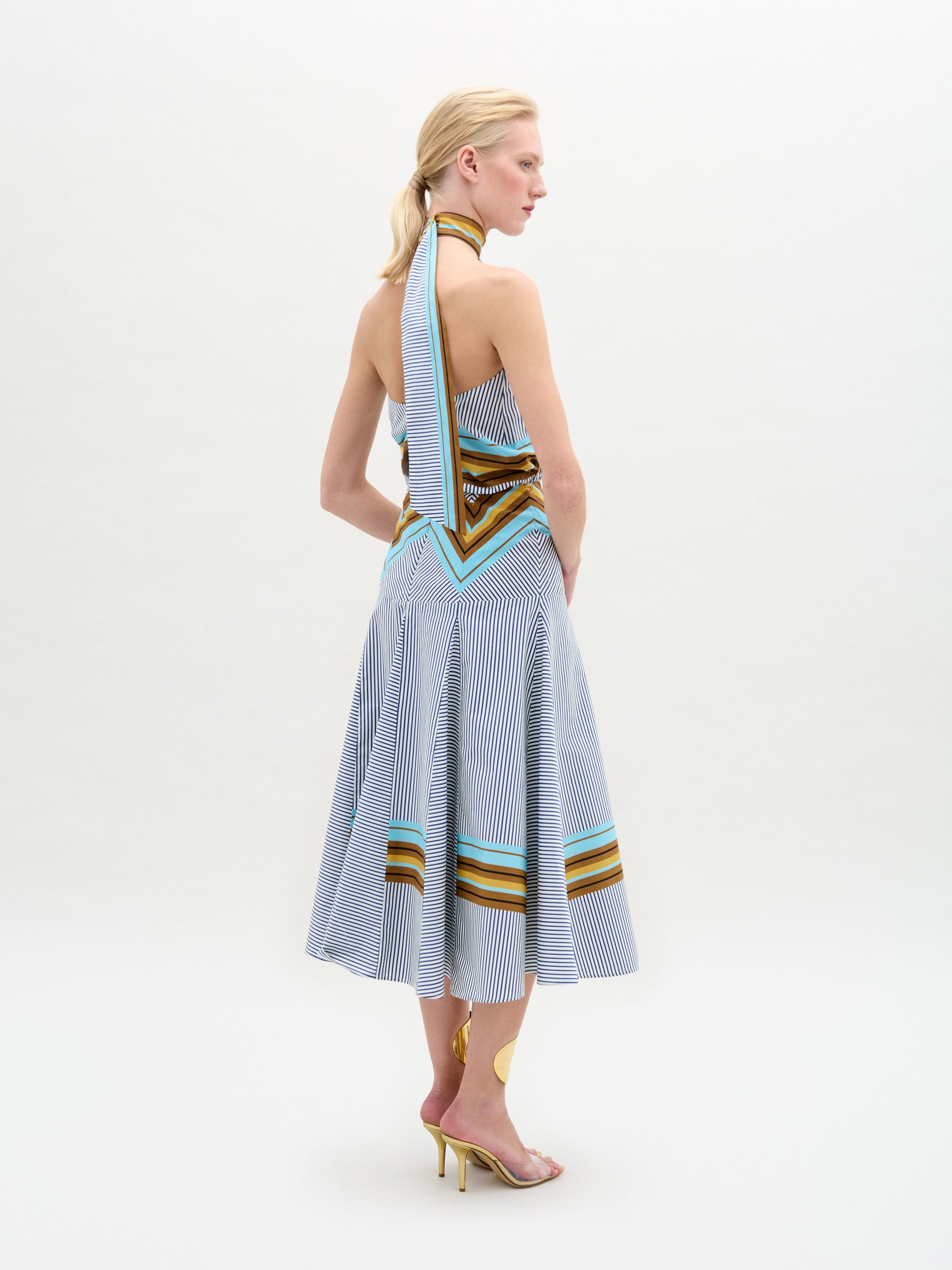 A person faces forward against a plain white background wearing the Elara Dress, a sleeveless, knee-length midi with geometric patterns and an A-line skirt, paired with ankle-strap heels.
