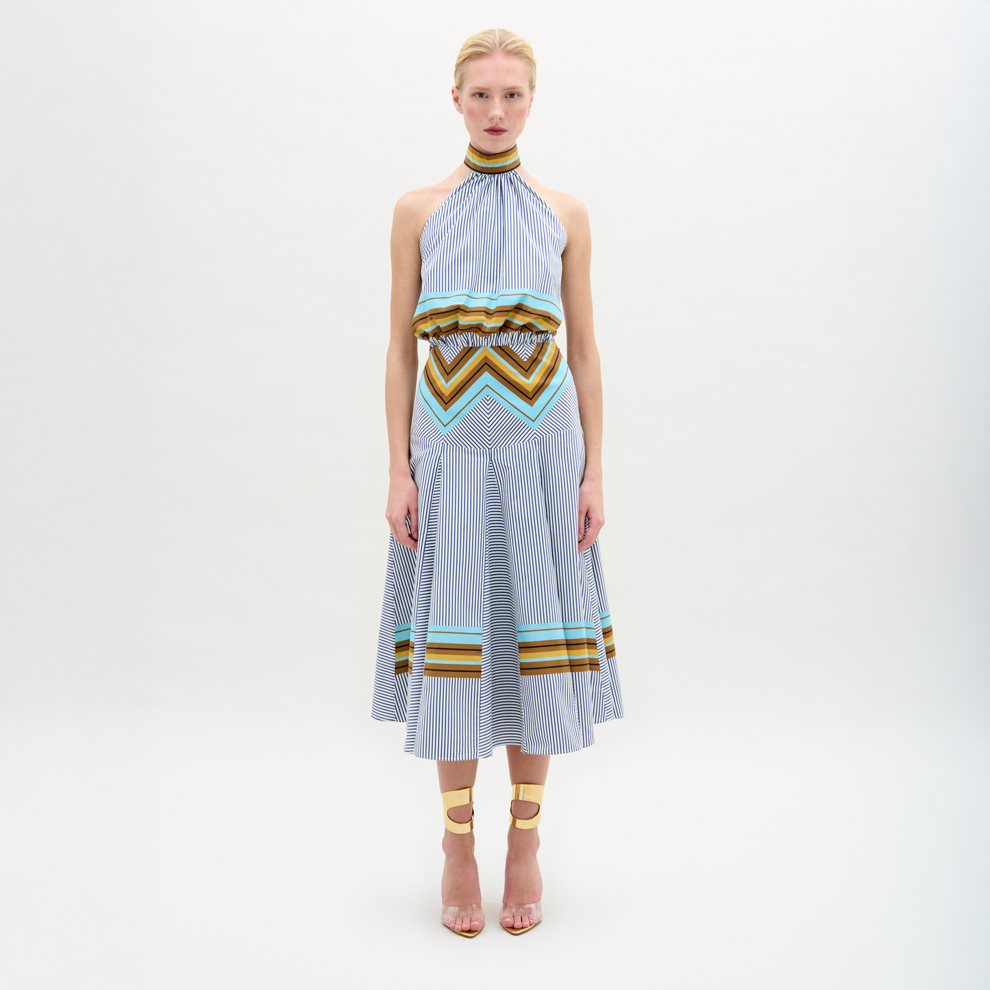 A person faces forward against a plain white background wearing the Elara Dress, a sleeveless, knee-length midi with geometric patterns and an A-line skirt, paired with ankle-strap heels.