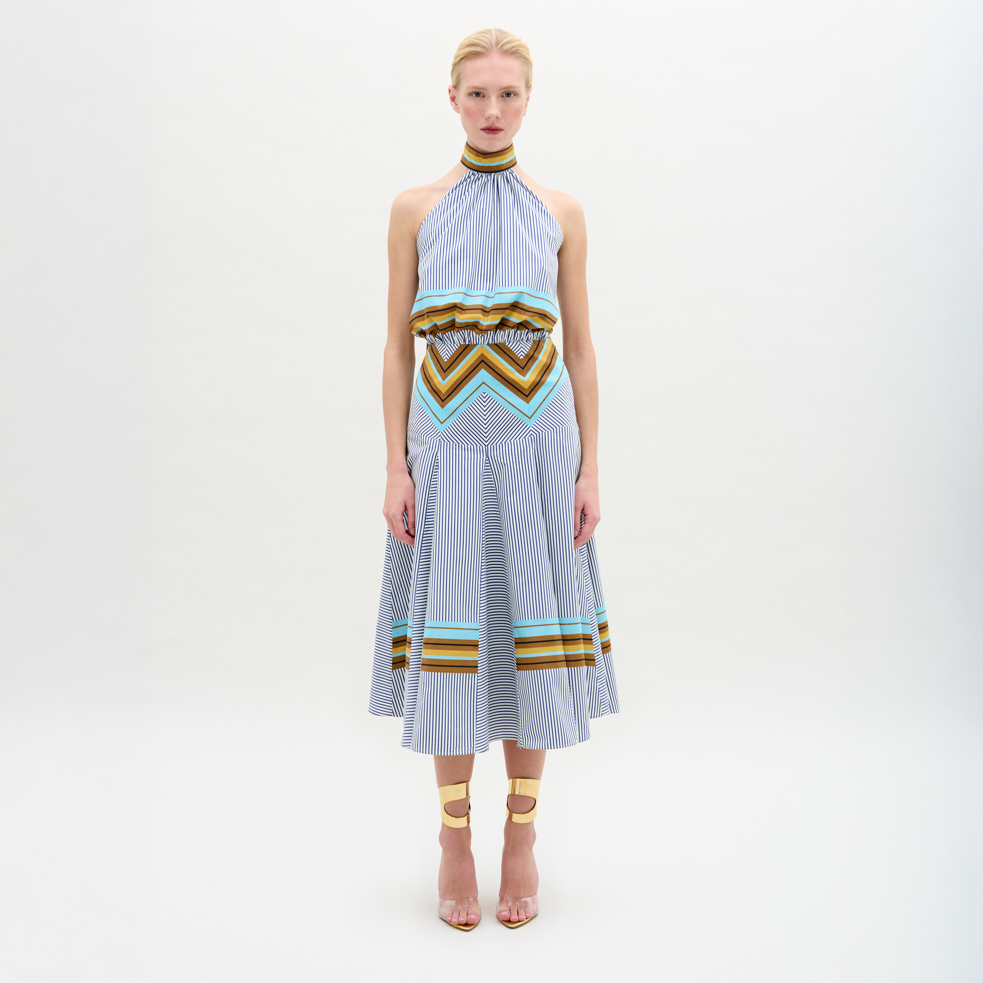 A person faces forward against a plain white background wearing the Elara Dress, a sleeveless, knee-length midi with geometric patterns and an A-line skirt, paired with ankle-strap heels.
