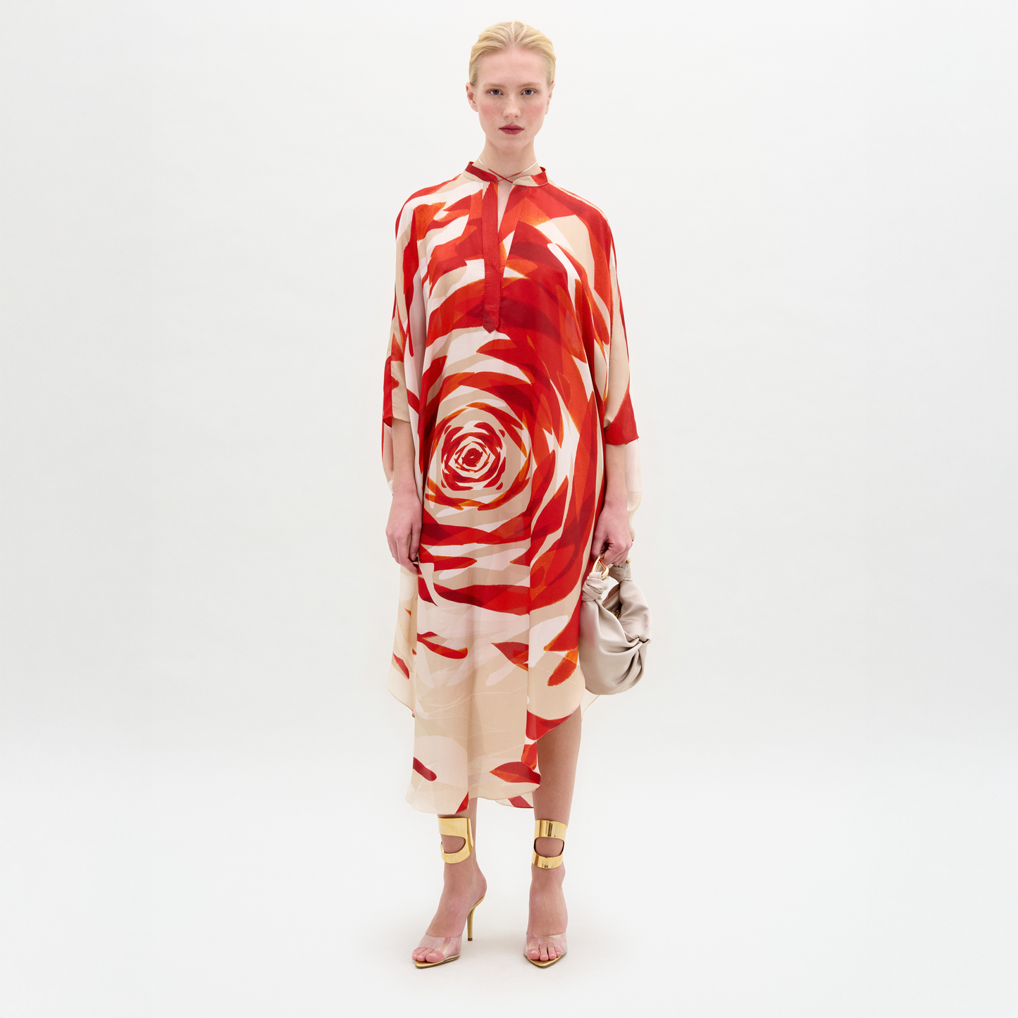 A woman stands against a white background wearing the Elea Tunic, a loose, knee-length dress made from Italian fabric with a red and cream spiral pattern. She holds a beige clutch and pairs the look with gold high-heeled sandals.