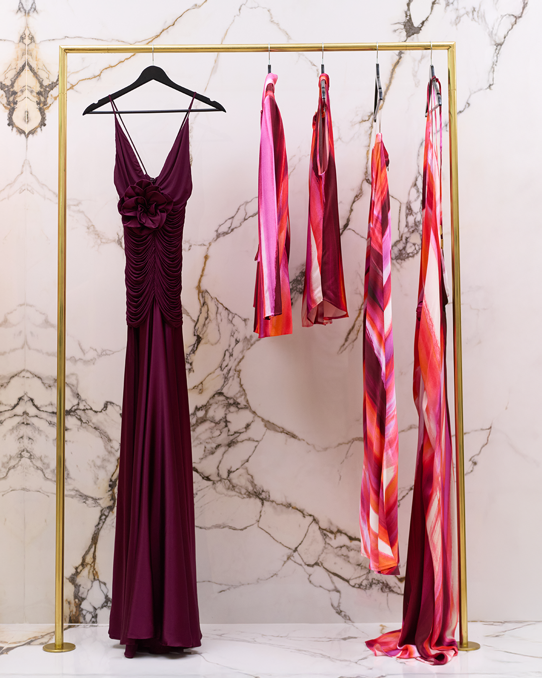 Elegant purple gown on a hanger with three pink and red patterned scarves hanging on a gold clothing rack against a marble wall.