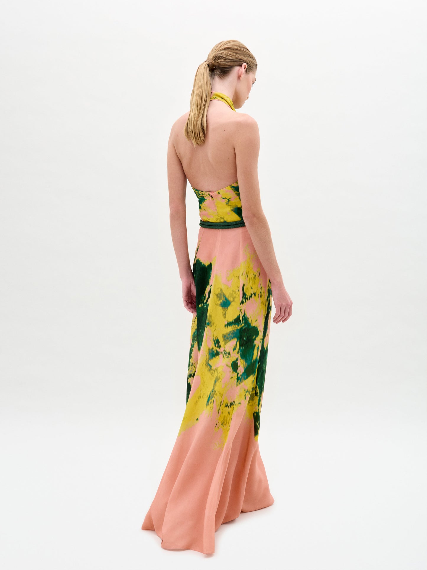 A person wearing the Ellis Dress, a floor-length, halter-neck style featuring green and yellow abstract patterns on a peach background, is looking to the side.