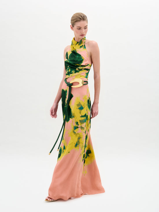 A person wearing the Ellis Dress, a floor-length, halter-neck style featuring green and yellow abstract patterns on a peach background, is looking to the side.