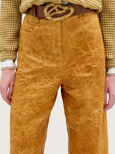 Person stands against a plain white background, wearing the Emine Pant—mustard yellow, high-waisted, wide-leg silhouette—paired with a white patterned button-up shirt and wide brown belt.