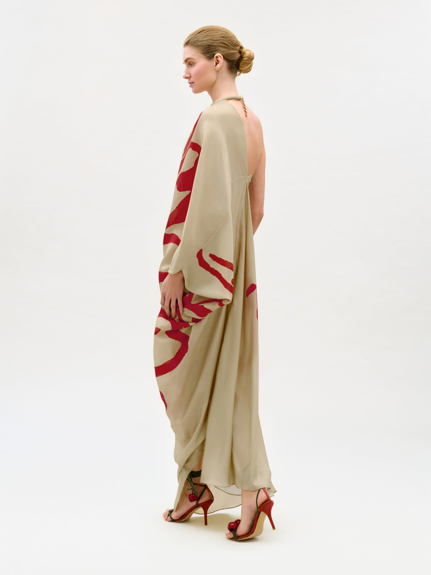 A woman in a low bun poses sideways in the Erica Dress, a beige, flowing silk maxi dress with bold red patterns and an open back. She pairs it with red high heels against a plain, light background.