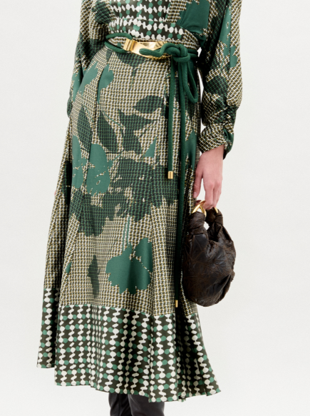 A woman stands against a white background wearing the Erin Skirt—a long, green and beige patterned skirt crafted from Italian fabric—paired with black heeled boots and a small black handbag.