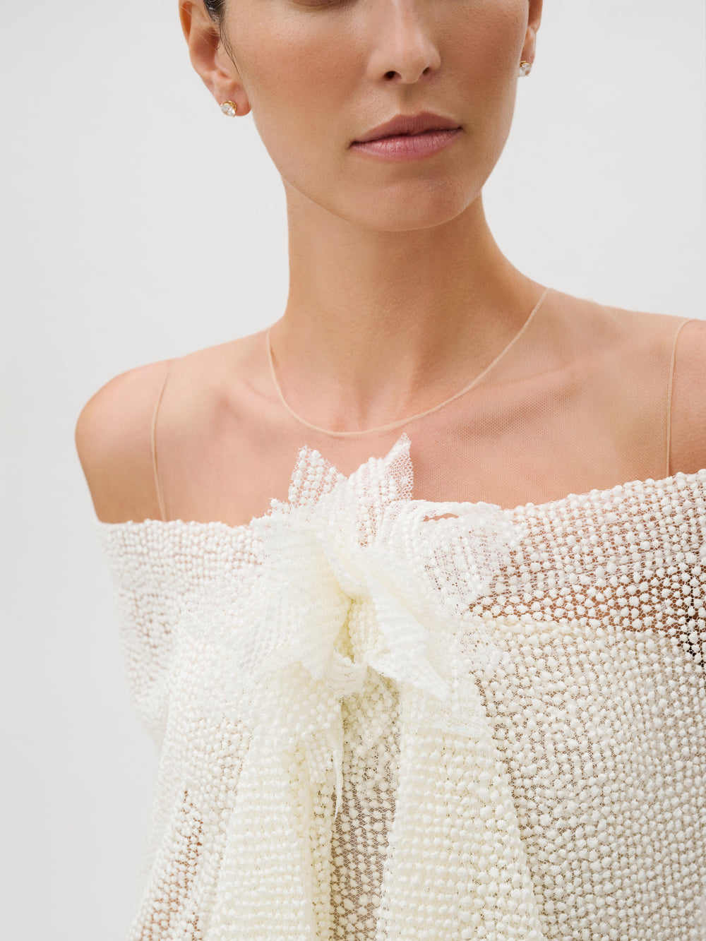 A woman wearing the Edna Dress—an off-shoulder, textured white piece featuring a large bow detail. She accessorizes with small earrings and a delicate necklace. The background is plain and light.