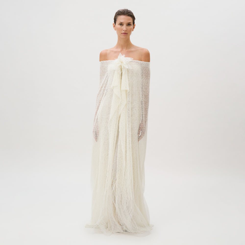 A woman stands against a plain white background, wearing the Edna Dress—an off-the-shoulder, textured white gown crafted from luxurious fabric with long, flowing layers and delicate sheer accents. This exquisite piece is masterfully made in Italy.