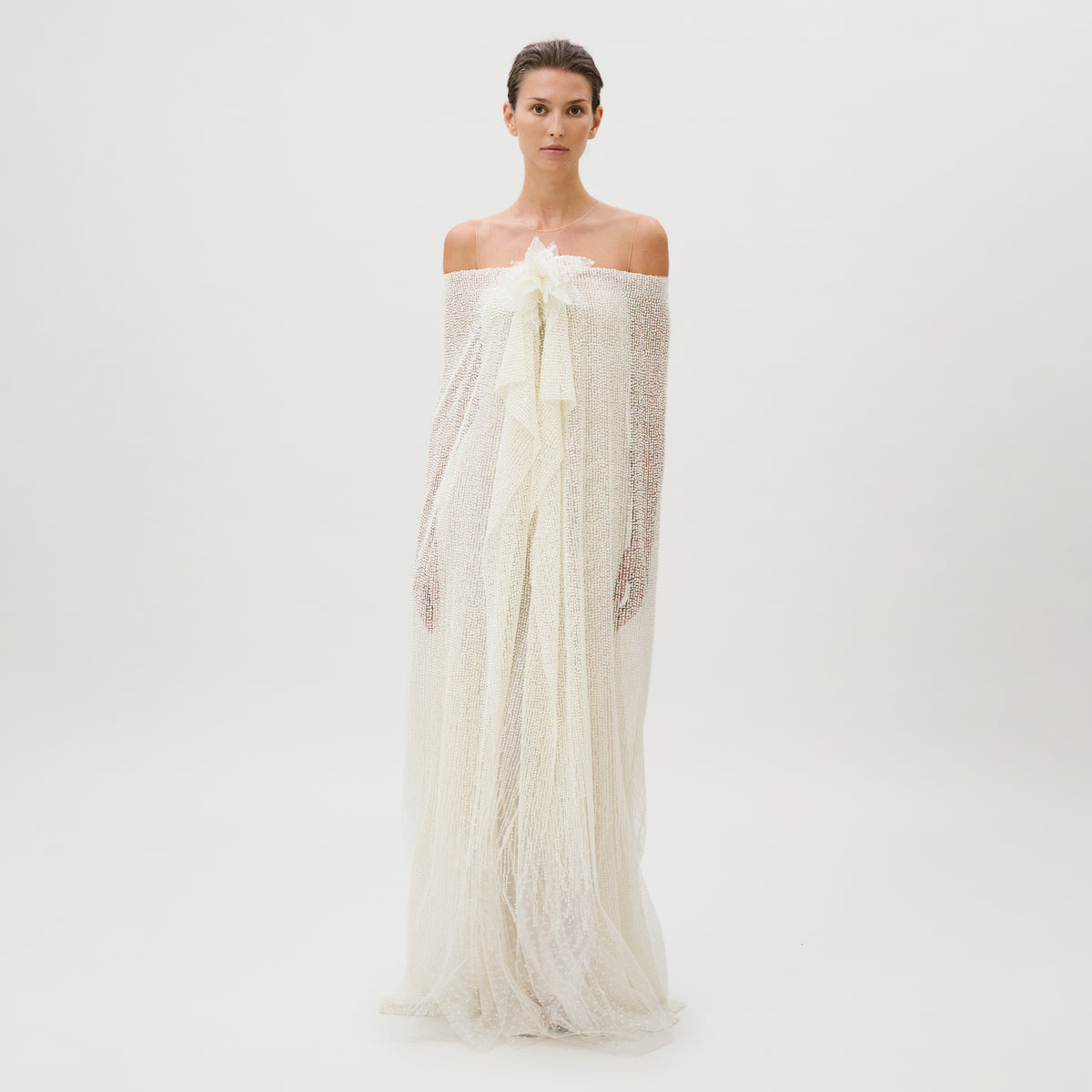 A woman stands against a plain white background, wearing the Edna Dress—an off-the-shoulder, textured white gown crafted from luxurious fabric with long, flowing layers and delicate sheer accents. This exquisite piece is masterfully made in Italy.