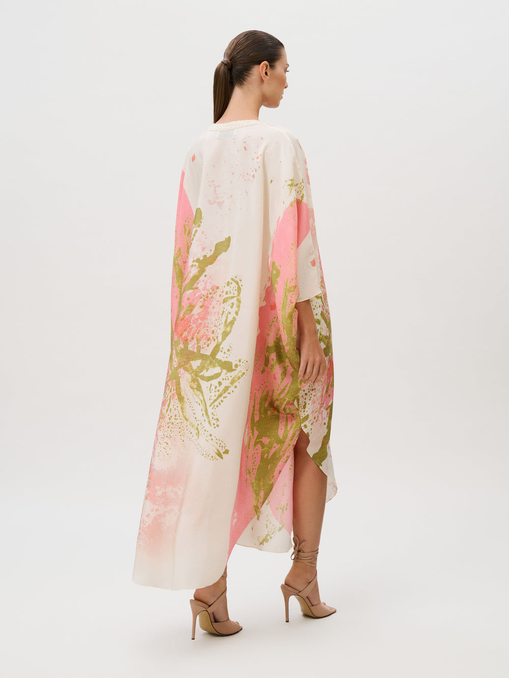 A person wears a loose, calf-length Elea Tunic in Italian silk with a pink and green abstract floral print, paired with lace-up high-heeled sandals.