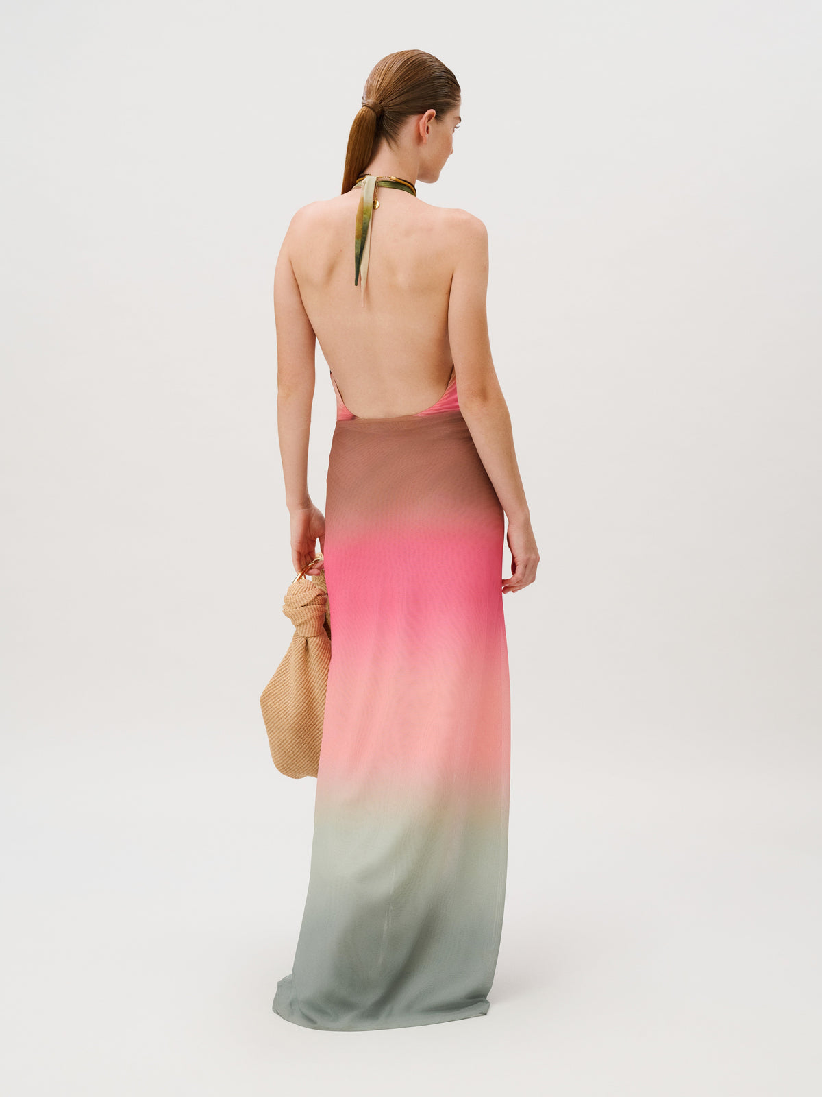 A woman wearing a slim fit, floor-length, backless Elif Skirt halter dress with a pink, brown, and green ombré gradient, holding a woven handbag.