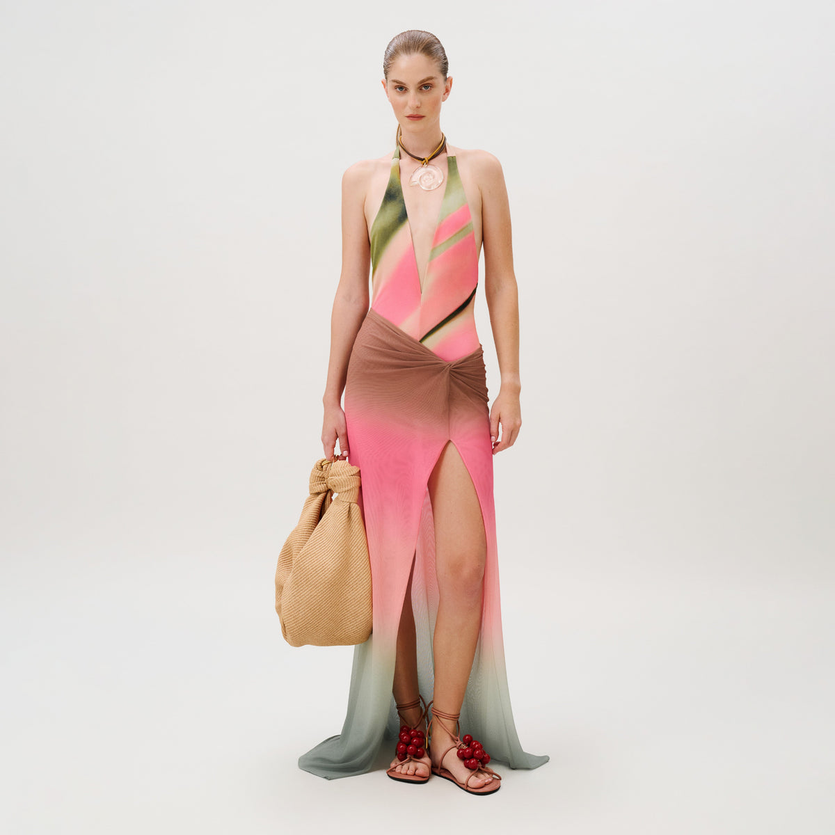 A model wears the Elif Skirt, a slim fit, floor-length, halter-neck dress with a green, pink, and brown gradient pattern, a knotted waist, and a high front slit, paired with a large straw bag and cherry-adorned flat sandals.