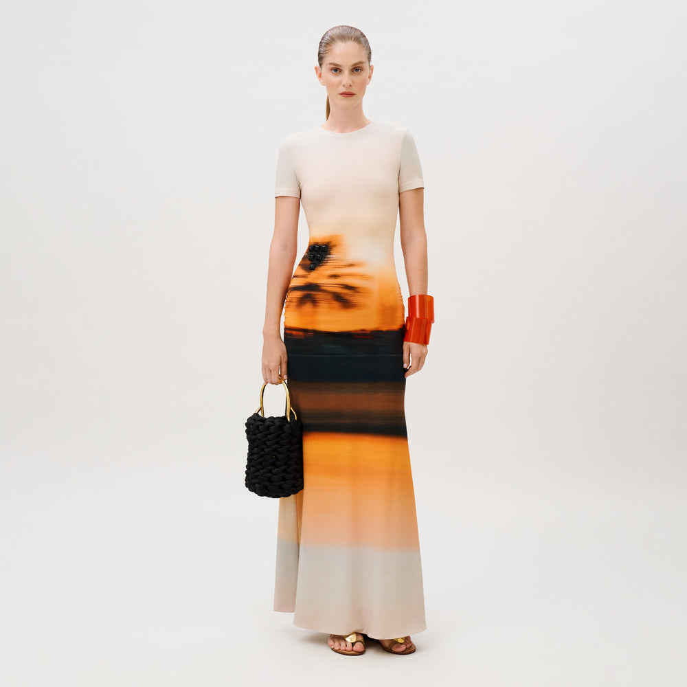A woman wears the Elora Dress, a slim-fit, floor-length viscose piece with an orange and black sunset-inspired gradient print, carrying a black woven handbag and wearing a thick orange bracelet.