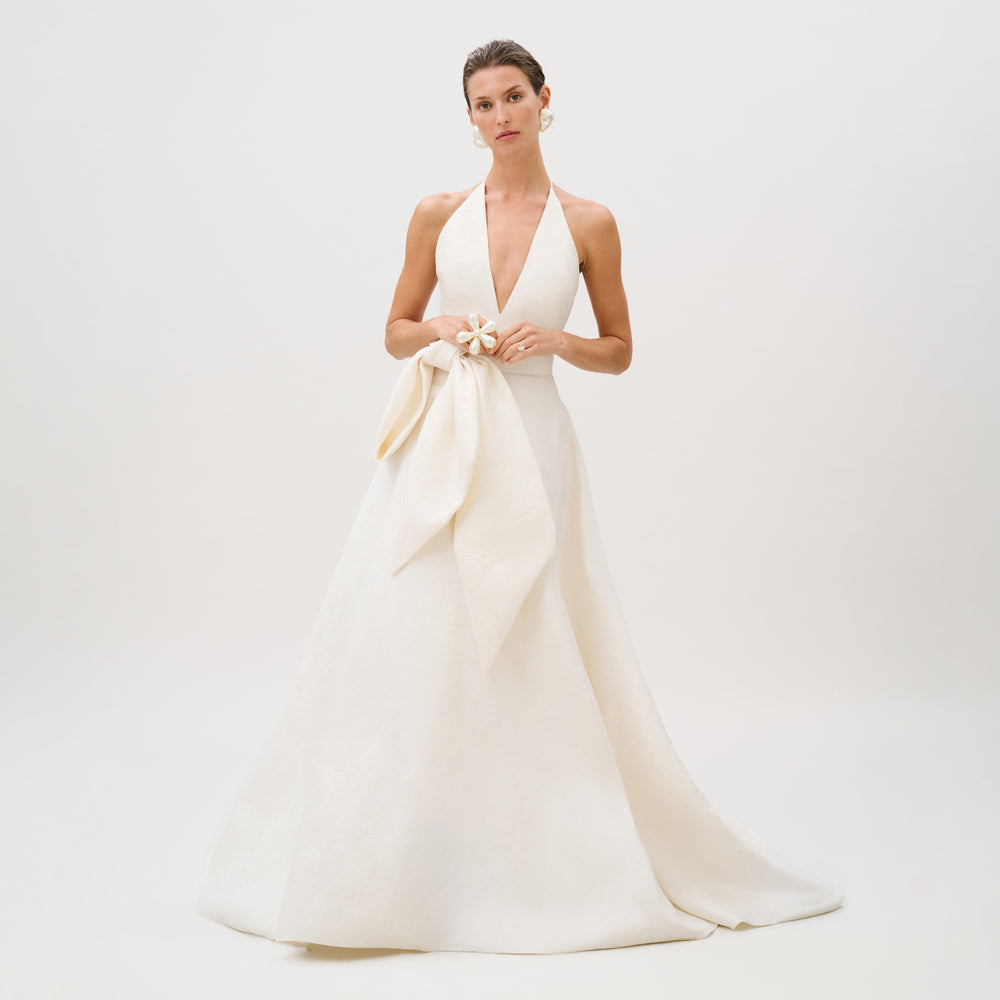 Dressed in the exquisite Elsa Dress—a sleeveless, floor-length white gown from our Atelier collection, crafted from luxurious Italian fabric and featuring a deep V neckline with a large bow at the waist—the woman stands against a plain light background.