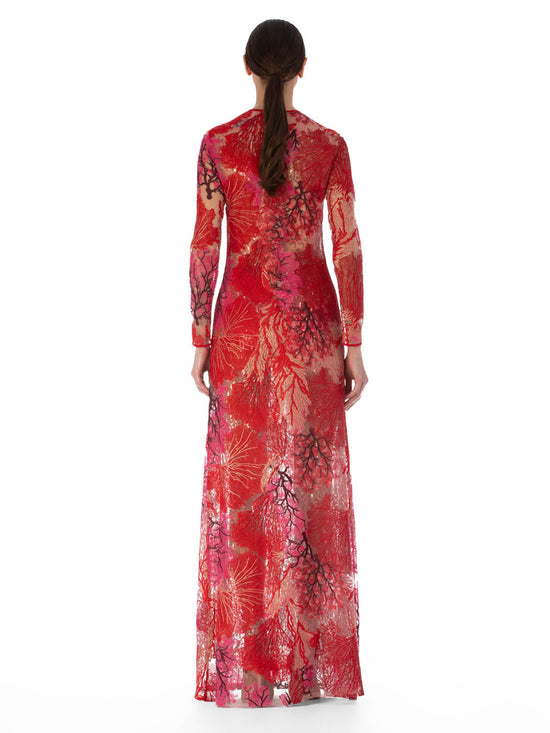 A person wearing the Enys Dress Multi Coral, a long maxi dress with intricate floral patterns and coral motifs, featuring a keyhole neckline, stands against a plain white background.