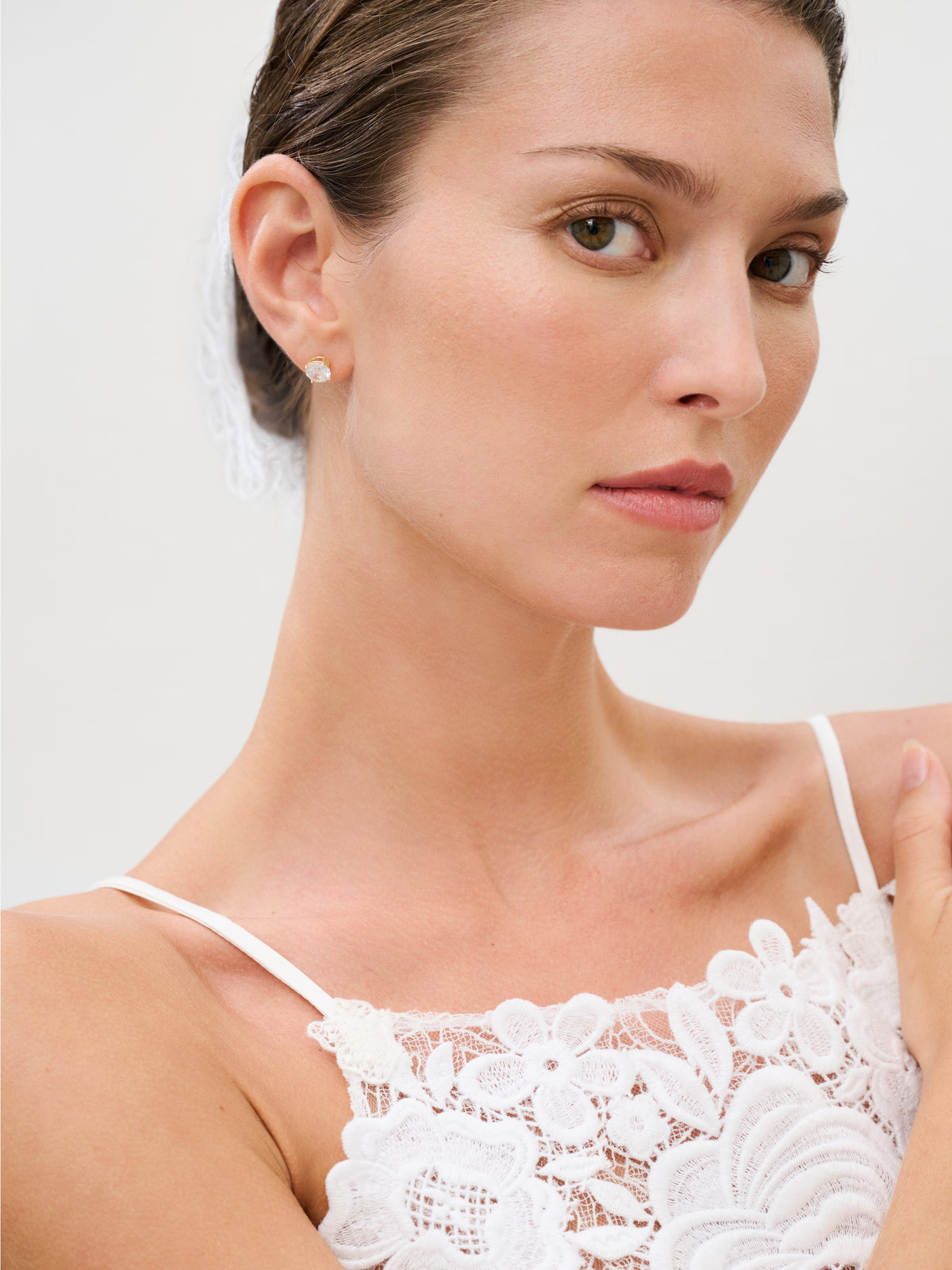 A woman with brown hair styled in an updo wears the Esme Dress, made from delicate white lace and Italian fabric, paired with small stud earrings. She gazes slightly over her shoulder with a neutral expression against a plain light backdrop. Made in Colombia.