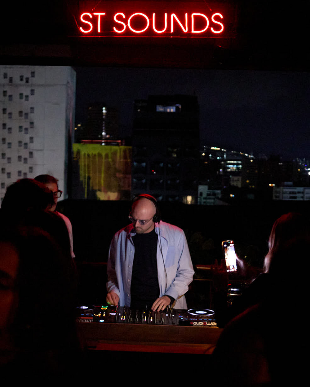 A DJ wearing a light blue jacket over a black shirt stands behind a mixing console at night, with a glowing red 'ST SOUNDS' neon sign above and a city skyline in the background.
