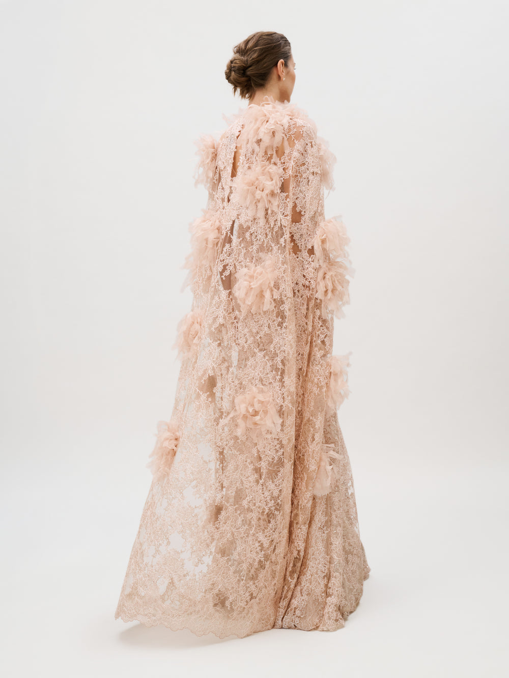 A woman in the Eudora Dress—a floor-length, pale pink lace gown adorned with floral appliqué and crafted from luxurious Italian fabric—stands against a plain white background, facing away from the camera with her hair styled in a low bun.