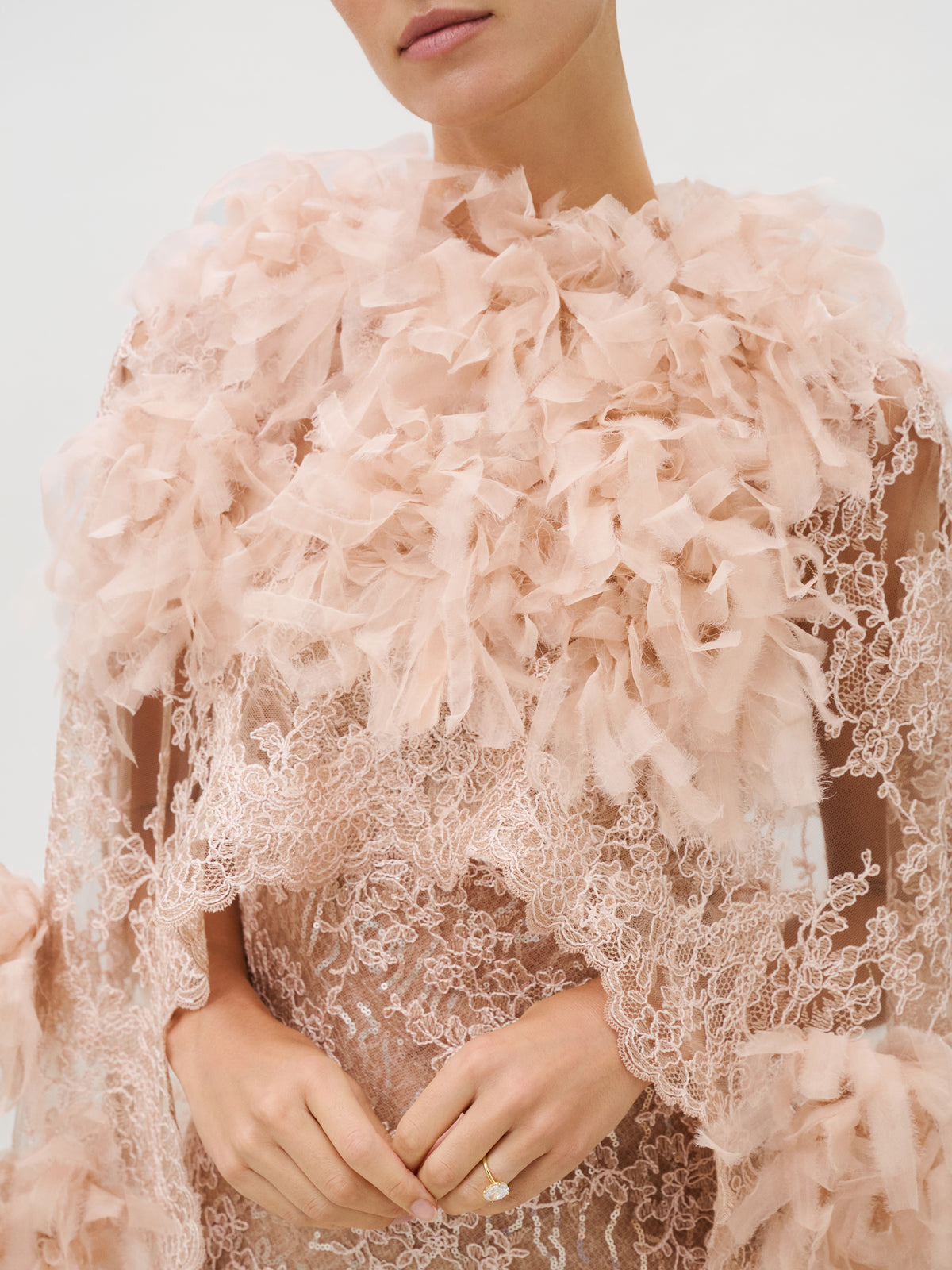 A person wearing the Eudora Dress—a delicate garment in pale pink, featuring exquisite Italian lace and soft, ruffled tulle accents around the shoulders and chest. The lower face is partially visible; one hand, adorned with a simple gold ring, rests gently by the waist.