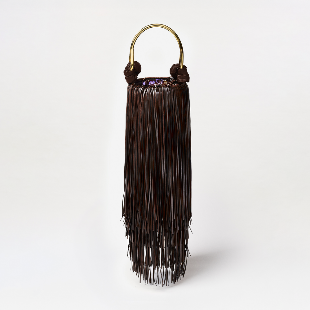 The Eugenia Bag is a dark brown eco leather handbag with long fringe strands and a gold semicircular handle, made in Colombia, displayed against a plain white background.