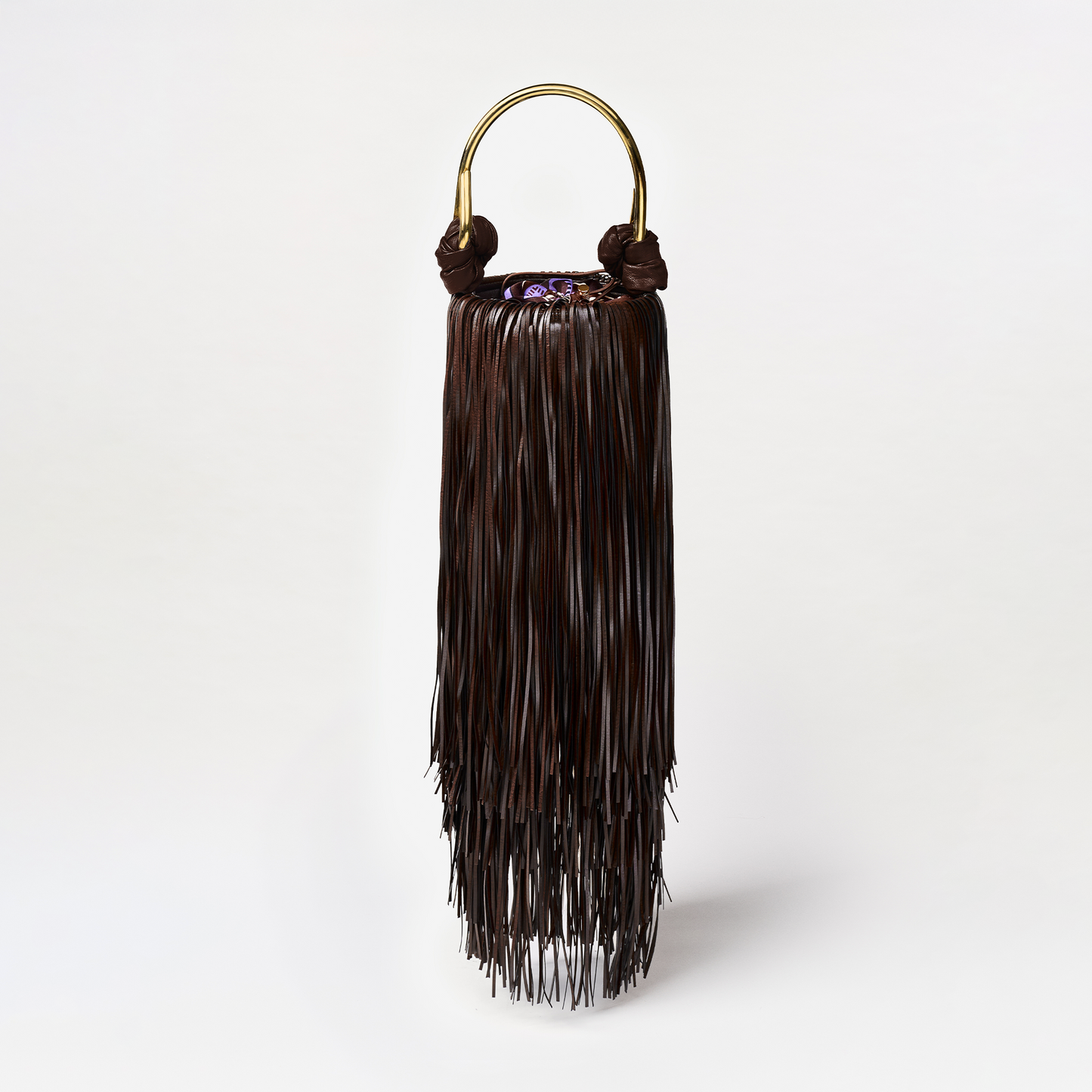 The Eugenia Bag is a dark brown eco leather handbag with long fringe strands and a gold semicircular handle, made in Colombia, displayed against a plain white background.