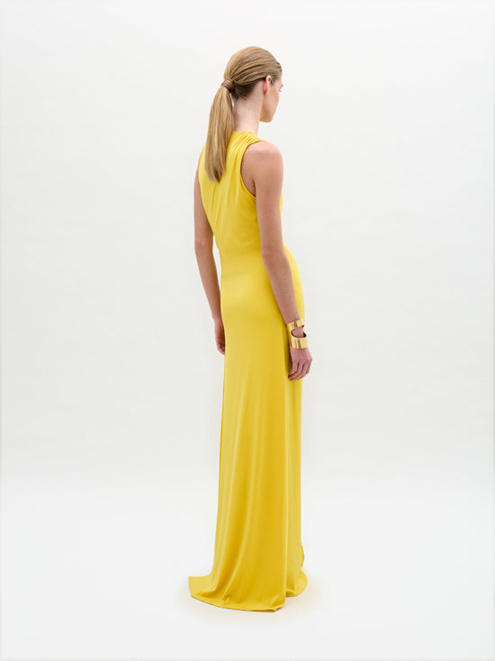 A woman stands against a plain background wearing the Fadila Dress, a long yellow gown featuring a sleeveless design, deep neckline, and a decorative circular clasp.