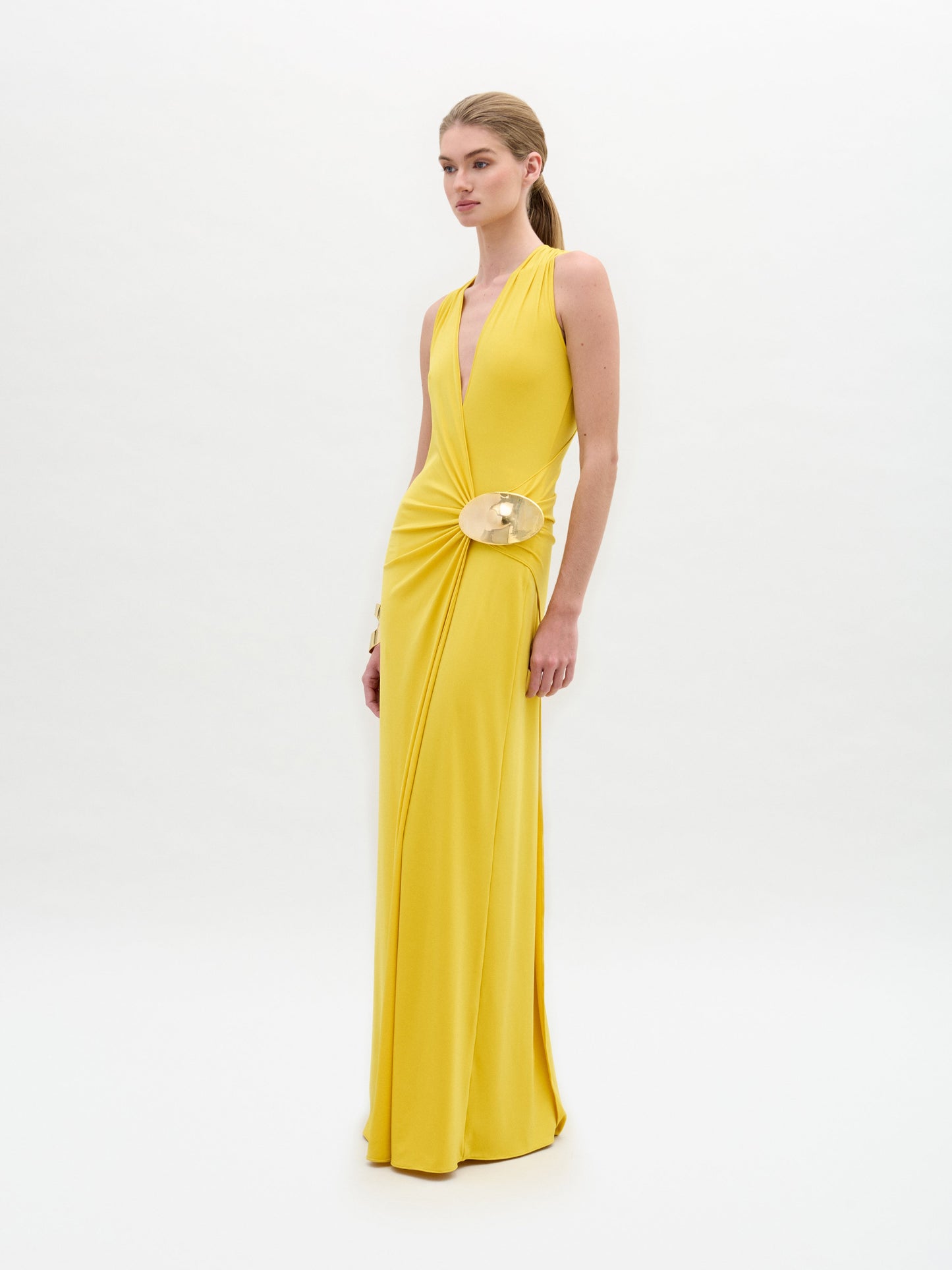 A woman stands against a plain background wearing the Fadila Dress, a long yellow gown featuring a sleeveless design, deep neckline, and a decorative circular clasp.