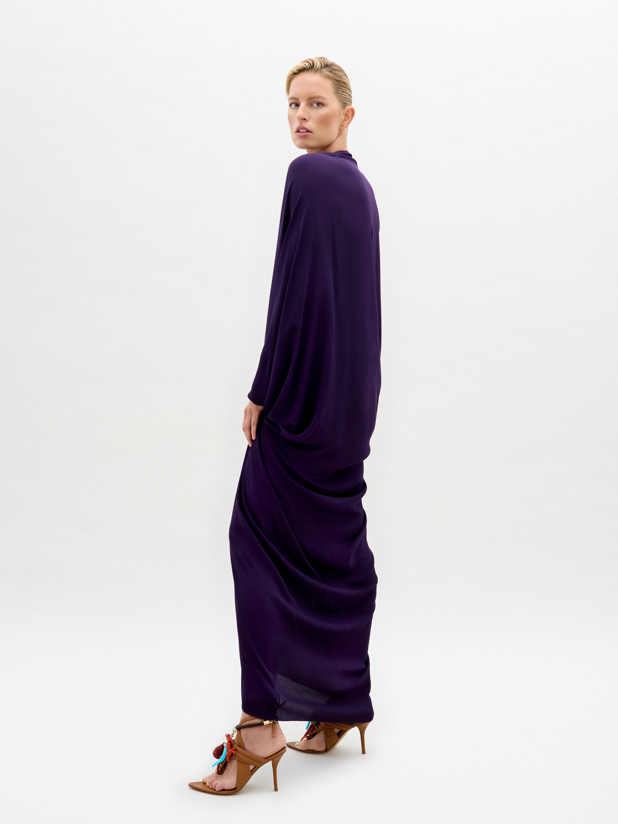 A person wearing a Fiore Tunic, a long dark purple gown with draped sleeves and a gathered waist, stands against a plain white background.