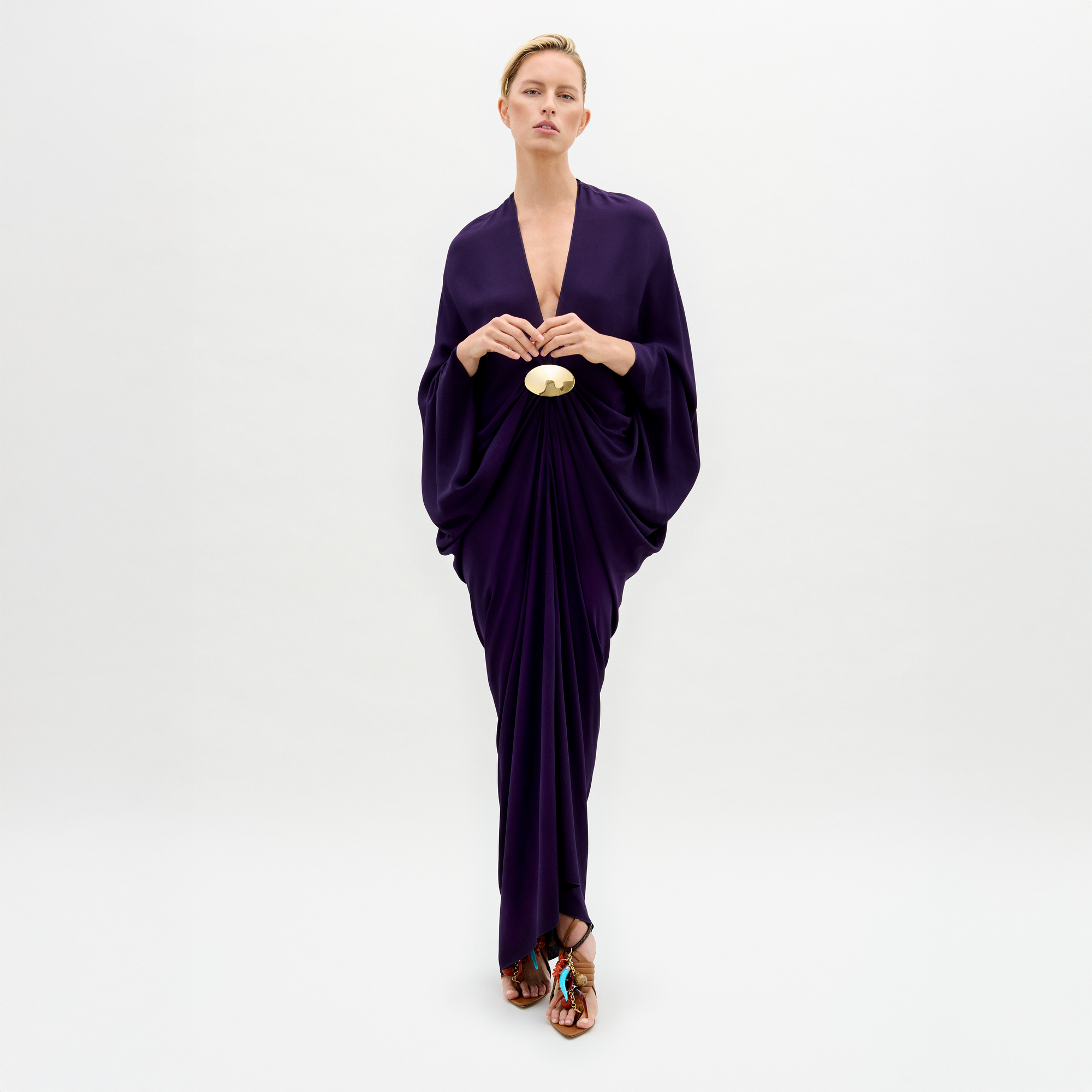 A person wearing a Fiore Tunic, a long dark purple gown with draped sleeves and a gathered waist, stands against a plain white background.