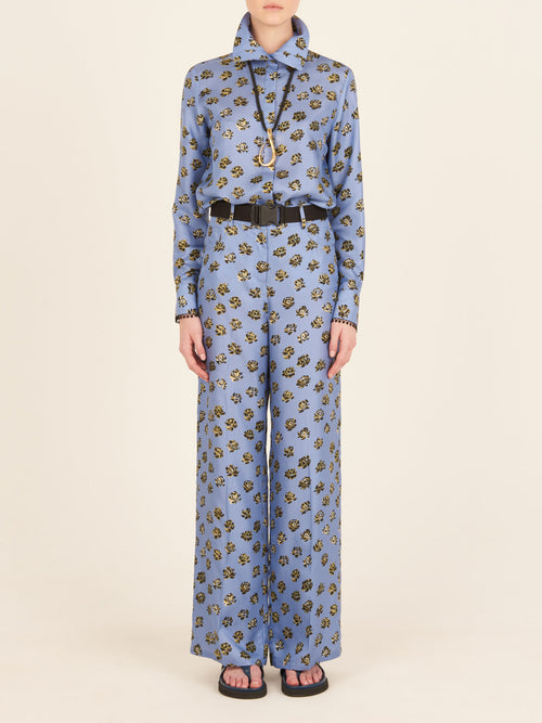 A person stands against a plain background, wearing the Emine Pant in Periwinkle, a blue outfit featuring flower patterns with long sleeves and high-waisted, wide-leg pants. The ensemble is completed with a black belt, necklace, and sandals.