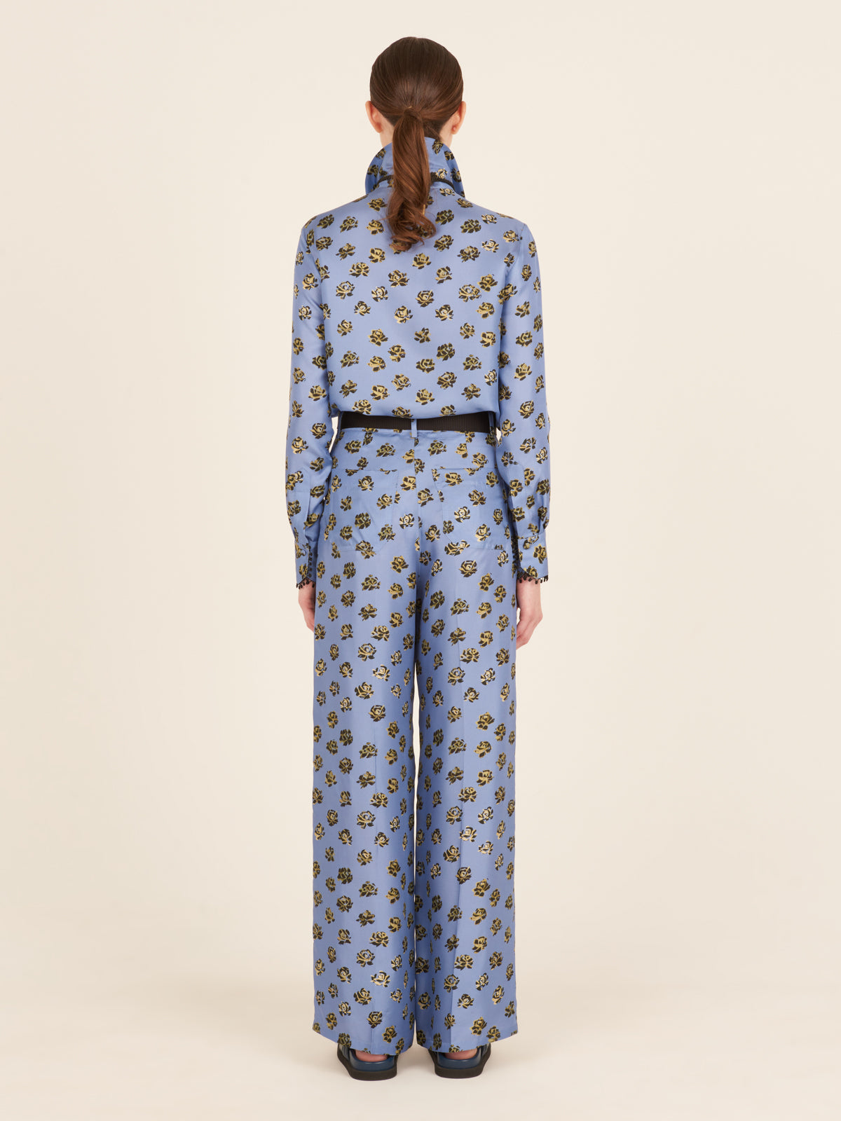 A person is depicted from the back wearing the Emine Pant in periwinkle, a blue floral-patterned outfit with a high collar and long sleeves, featuring wide-legged printed trousers.