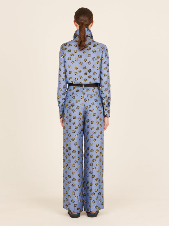 A person is depicted from the back wearing the Emine Pant in periwinkle, a blue floral-patterned outfit with a high collar and long sleeves, featuring wide-legged printed trousers.