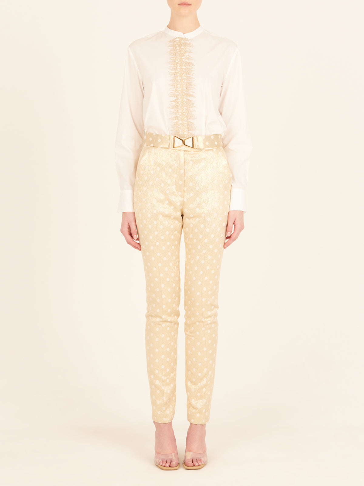 Rear view of a person with long hair in a ponytail, wearing a white blouse and the Orion Pant Gold Inverted, featuring high-waisted trousers with a straight leg silhouette, standing in heels on a plain background.