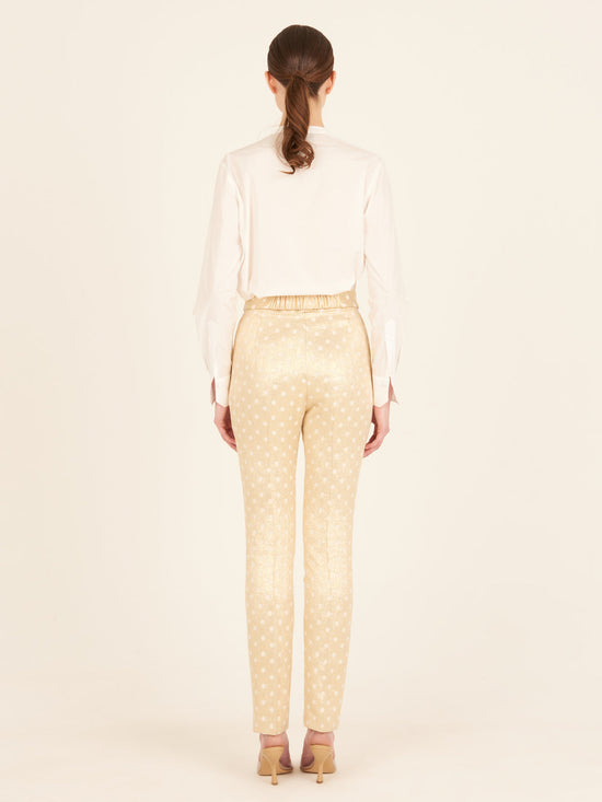 Rear view of a person with long hair in a ponytail, wearing a white blouse and the Orion Pant Gold Inverted, featuring high-waisted trousers with a straight leg silhouette, standing in heels on a plain background.