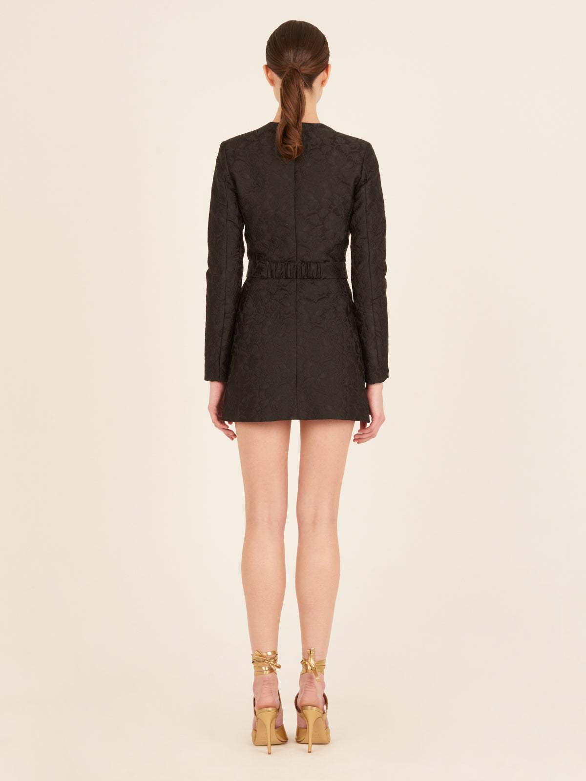 Romana Dress Black: A black long-sleeve mini dress featuring a floral pattern, front zipper, and gold bow belt, crafted from brocade fabric for an elegant touch.