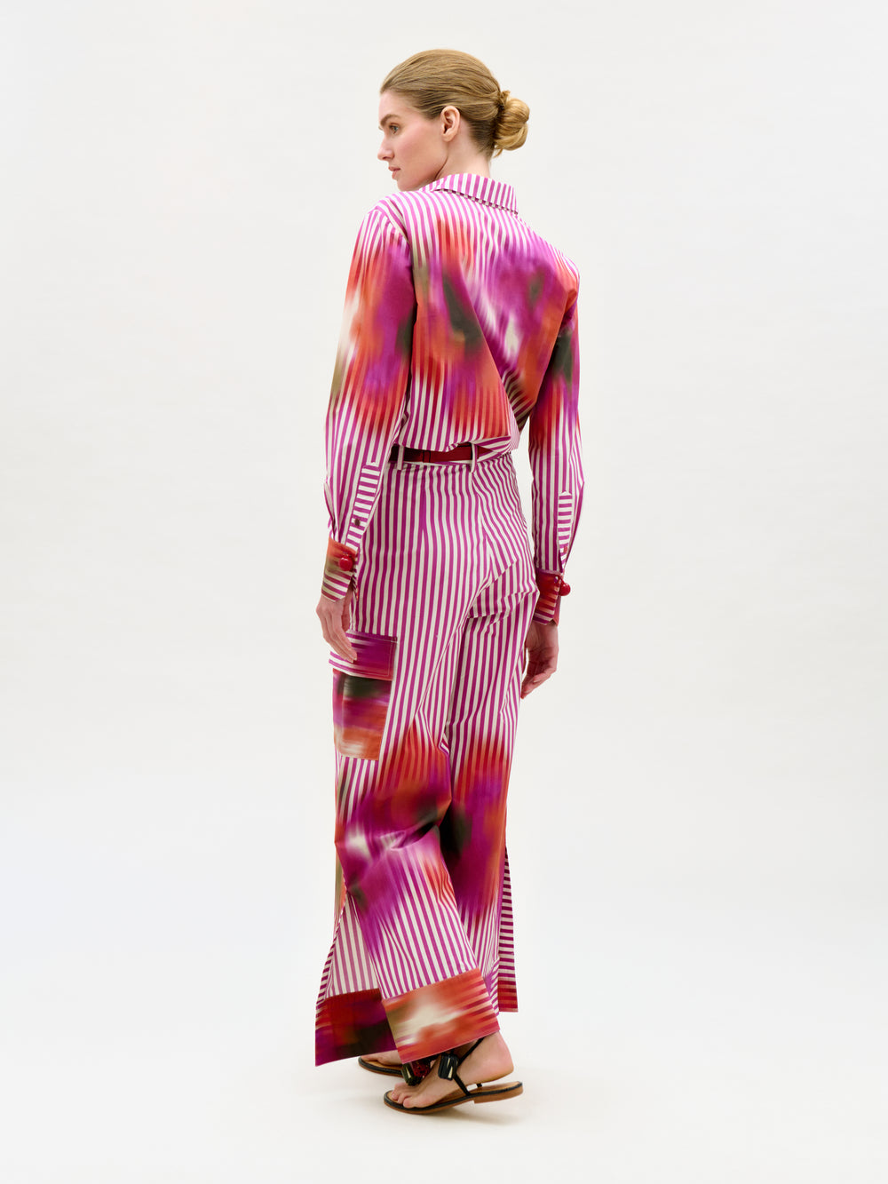 A woman faces away, wearing the Falak Pant—crafted from luxurious Italian fabric with magenta, red, and white stripes and splashes. Her hair is in a neat bun, and she wears black sandals.