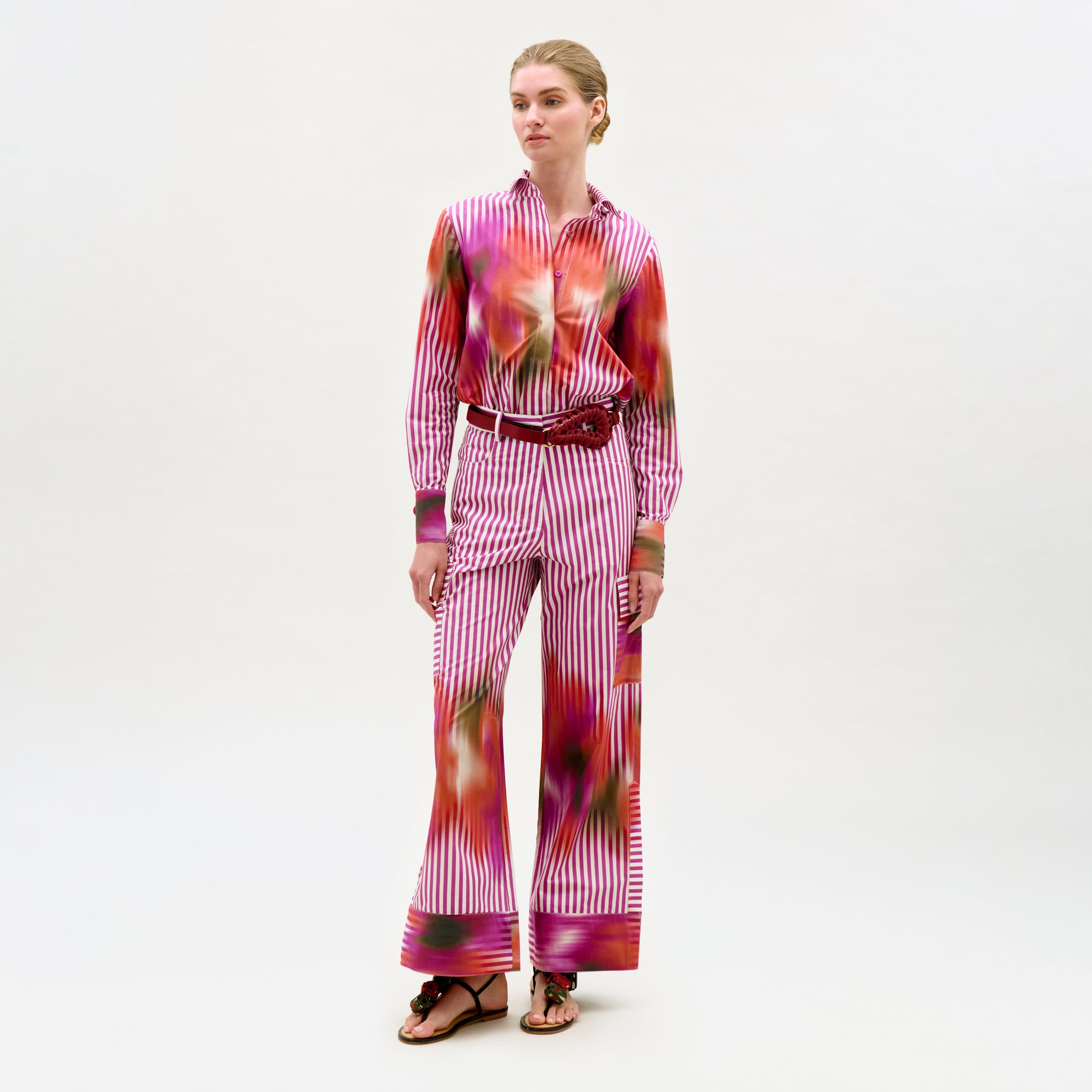 A woman wears the Falak Pant—a high-rise, straight leg style in pink, red, and white tones—paired with a colorful patterned blouse, red belt, and sandals against a plain white background.
