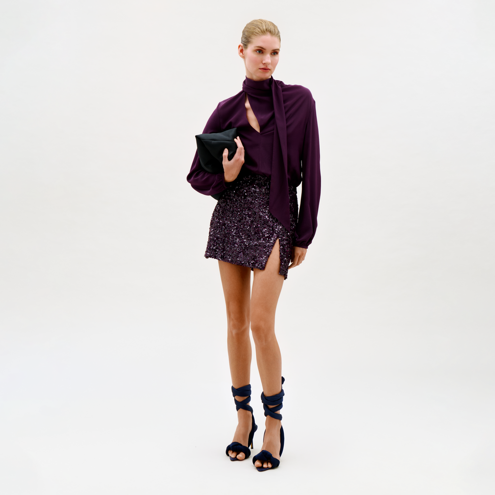 A woman stands against a plain background, wearing the Faye Dress, styled with a black clutch and high-heeled sandals featuring wide ankle straps.